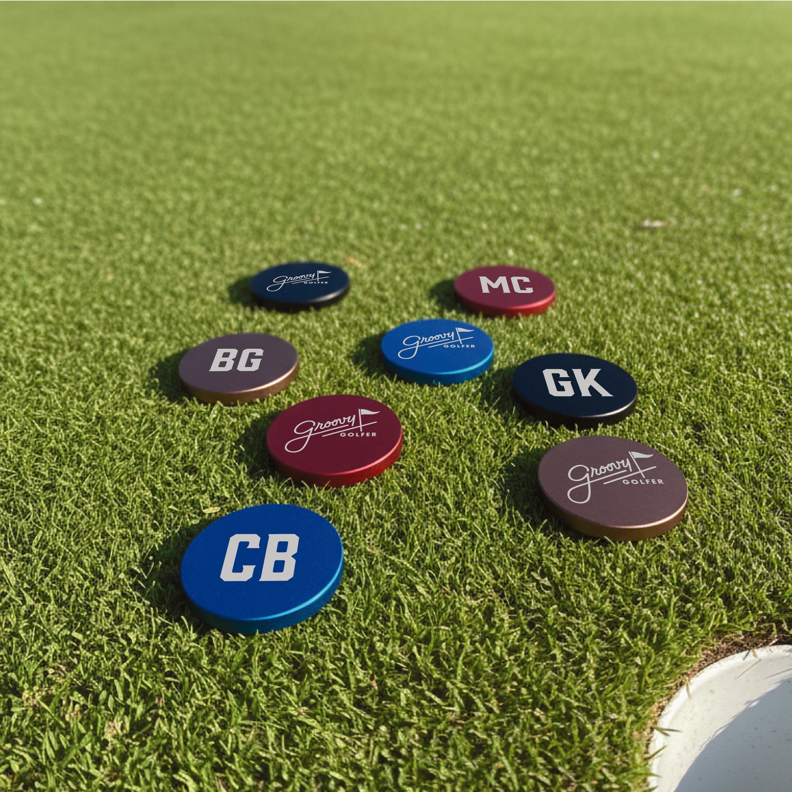 Seven oval ball markers displayed on green golf course grass, showing sample personalization with initials including BG, MC, GK, CB and Groovy Golfer logo designs in navy, maroon, and blue finishes