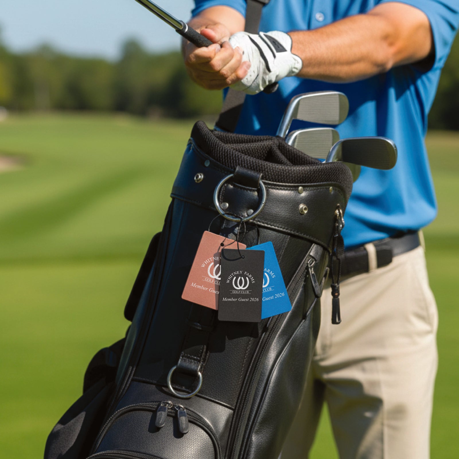 Golfer holding club with black leather golf bag displaying three sample bag tags in tan, black, and blue, attached via steel cable loops, on a course green background