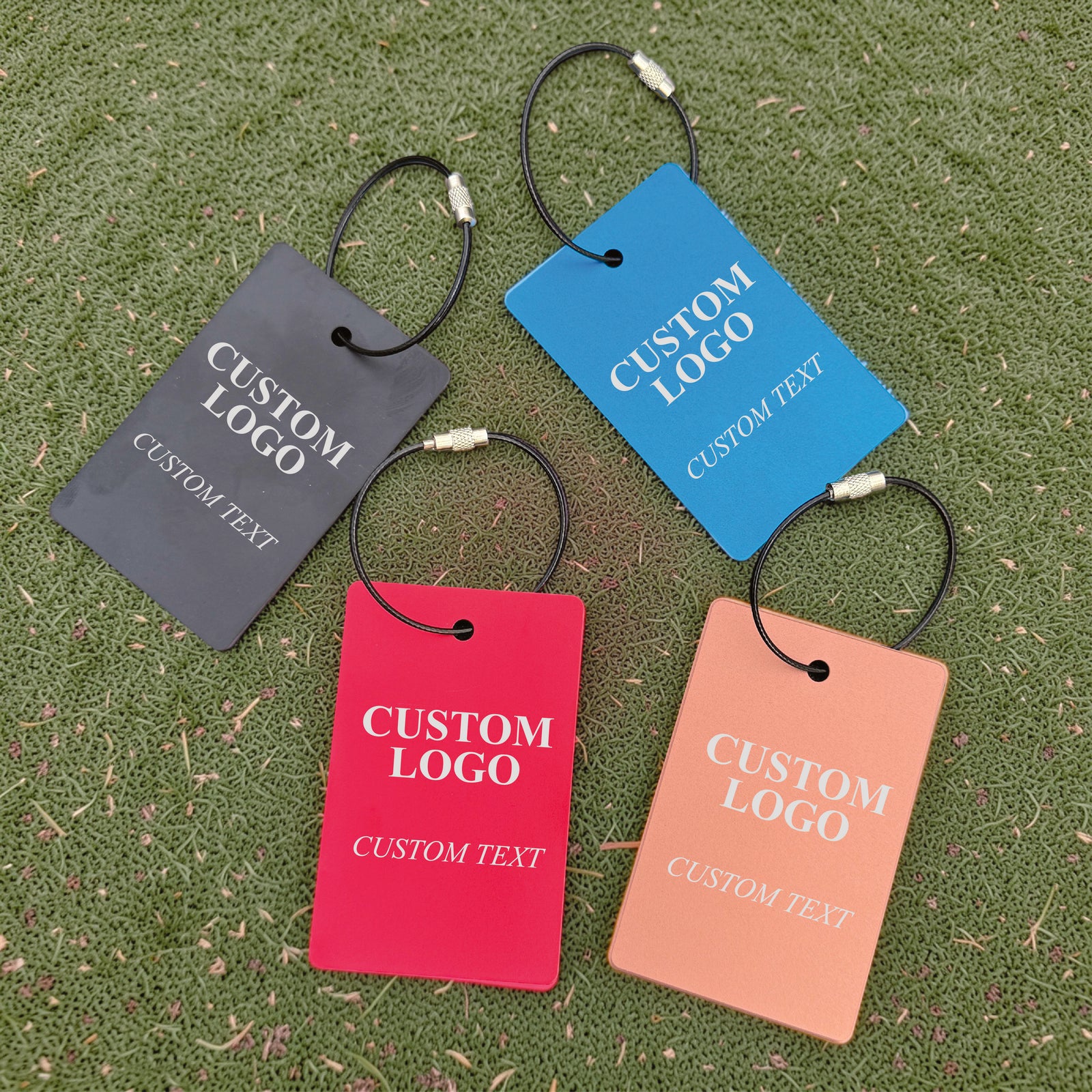Four personalized golf bag tags in charcoal, blue, red, and peach with sample logos and text, displayed on grass with steel cable attachments