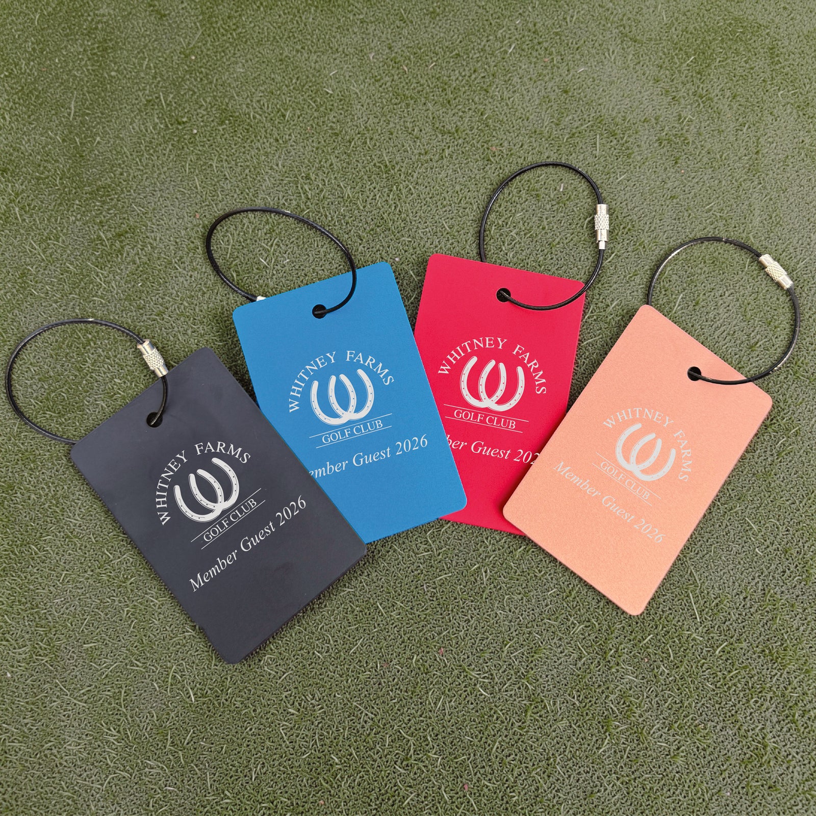 Four custom golf bag tags in charcoal, blue, red, and peach, each engraved with Whitney Farms Golf Club logo and sample text "Member Guest 2026", displayed on artificial turf with black cable loops.