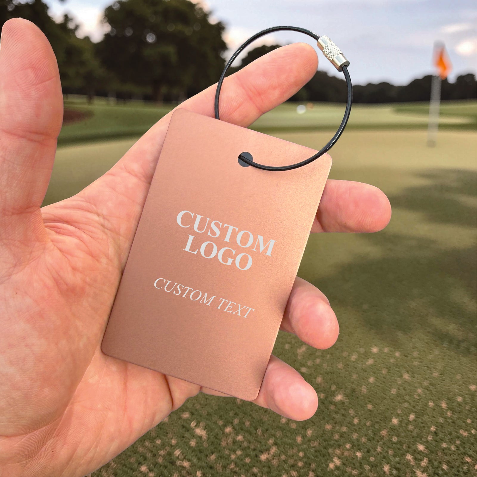 Rose gold aluminum golf bag tag held in hand on golf course, engraved with sample "CUSTOM LOGO" and "CUSTOM TEXT" in white lettering, with black steel cable loop attachment