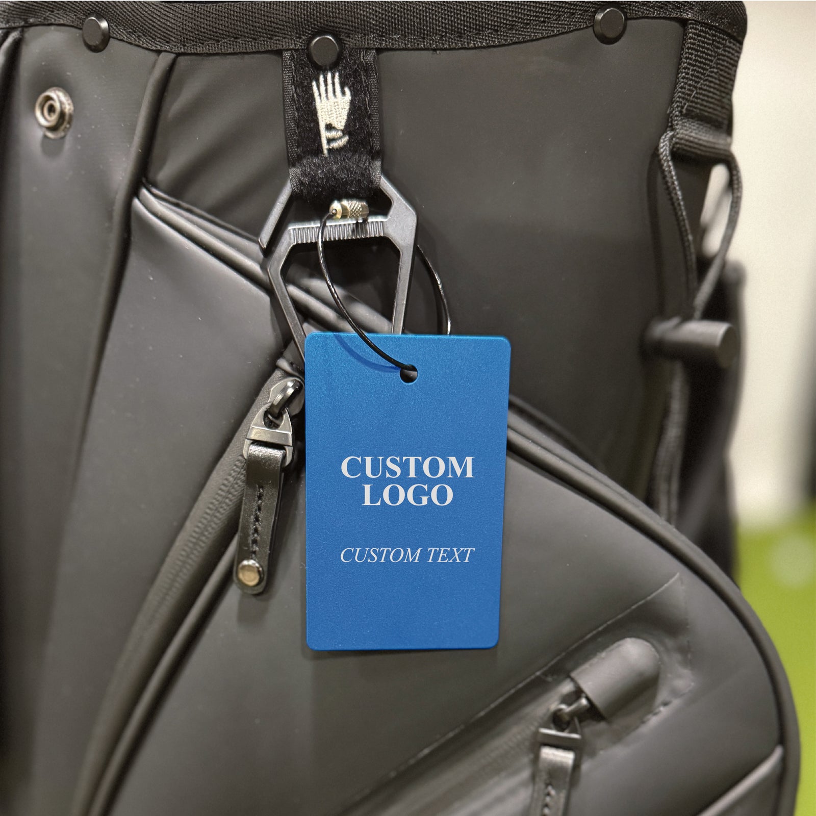 Blue aluminum golf bag tag with black cable loop attached to golf bag, showing sample custom logo and text placeholder areas