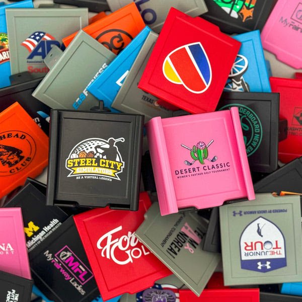 Collection of custom logo phone caddies in various colors including black, pink, red, and gray, displayed with sample golf tournament logos printed on the faceplates.