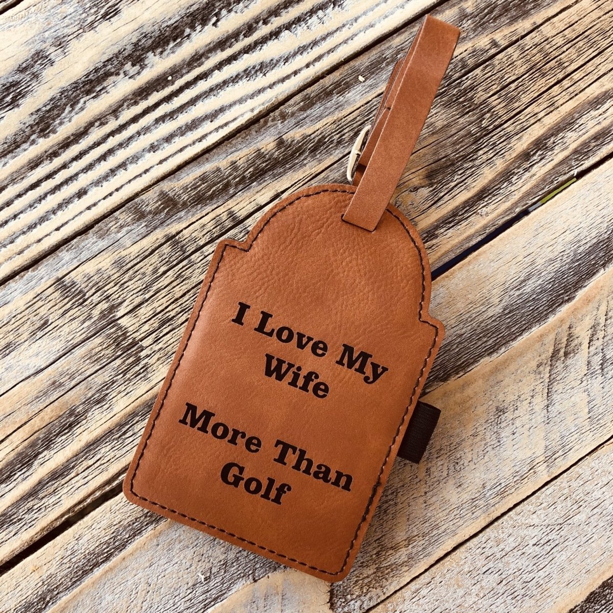 Tan leather golf bag tag with engraved sample text "I Love My Wife More Than Golf" and brown stitched edges, displayed on wooden surface