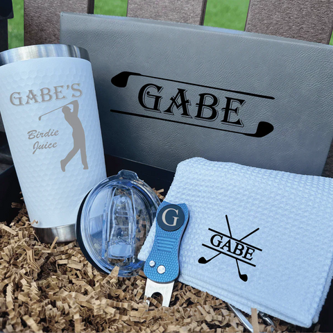 Assortment of personalized golf gifts under $100 including an engraved decanter, custom golf towel, monogrammed shoe bag and a magnetic cart koozie arranged on turf