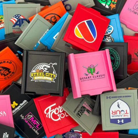 Assorted custom golf towels and accessories with sample logos and text in various colors including red, pink, black, gray, and blue.