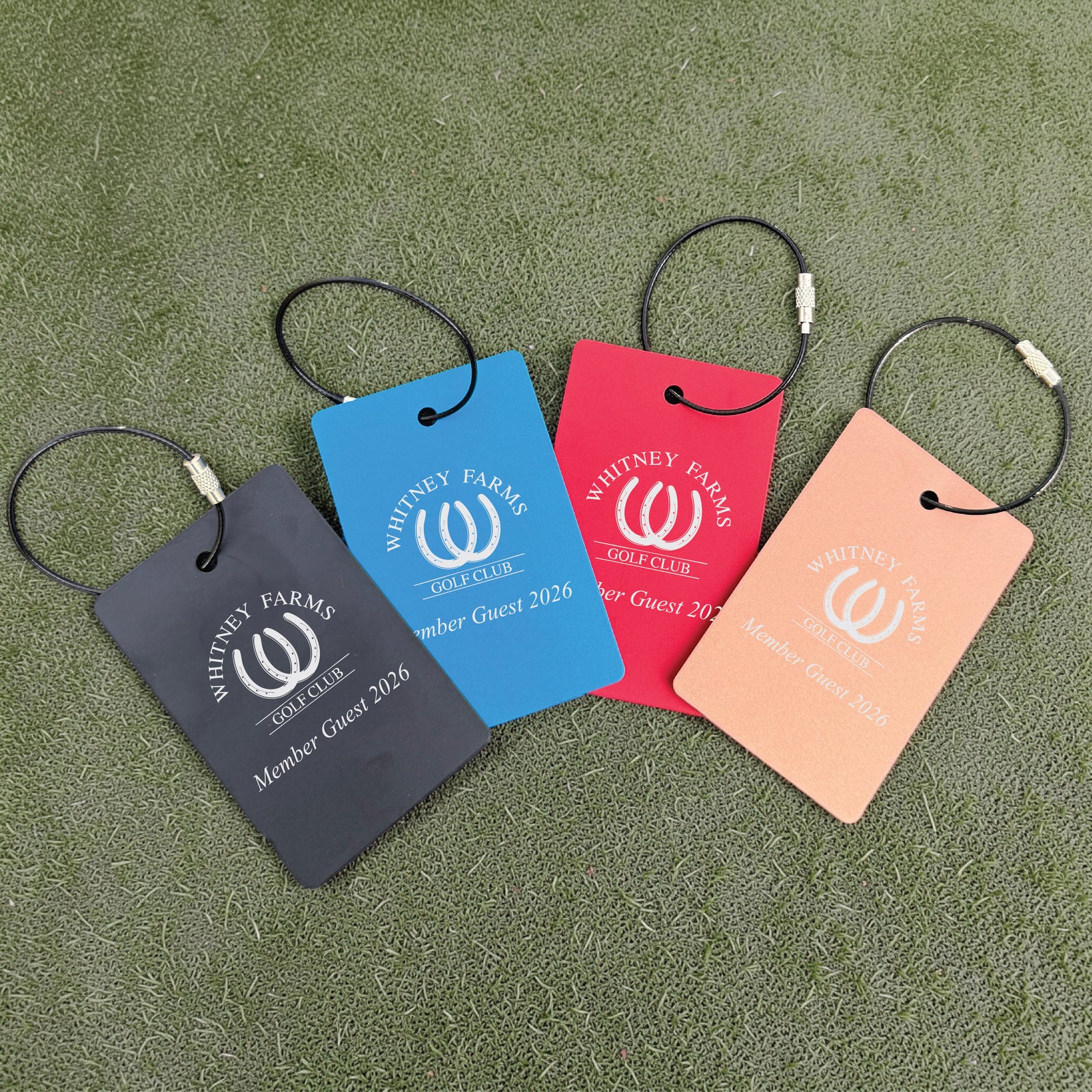 Four golf bag tags in charcoal, blue, red, and peach, each engraved with sample Whitney Farms Golf Club logo and "Member Guest 2026" text, displayed on green turf