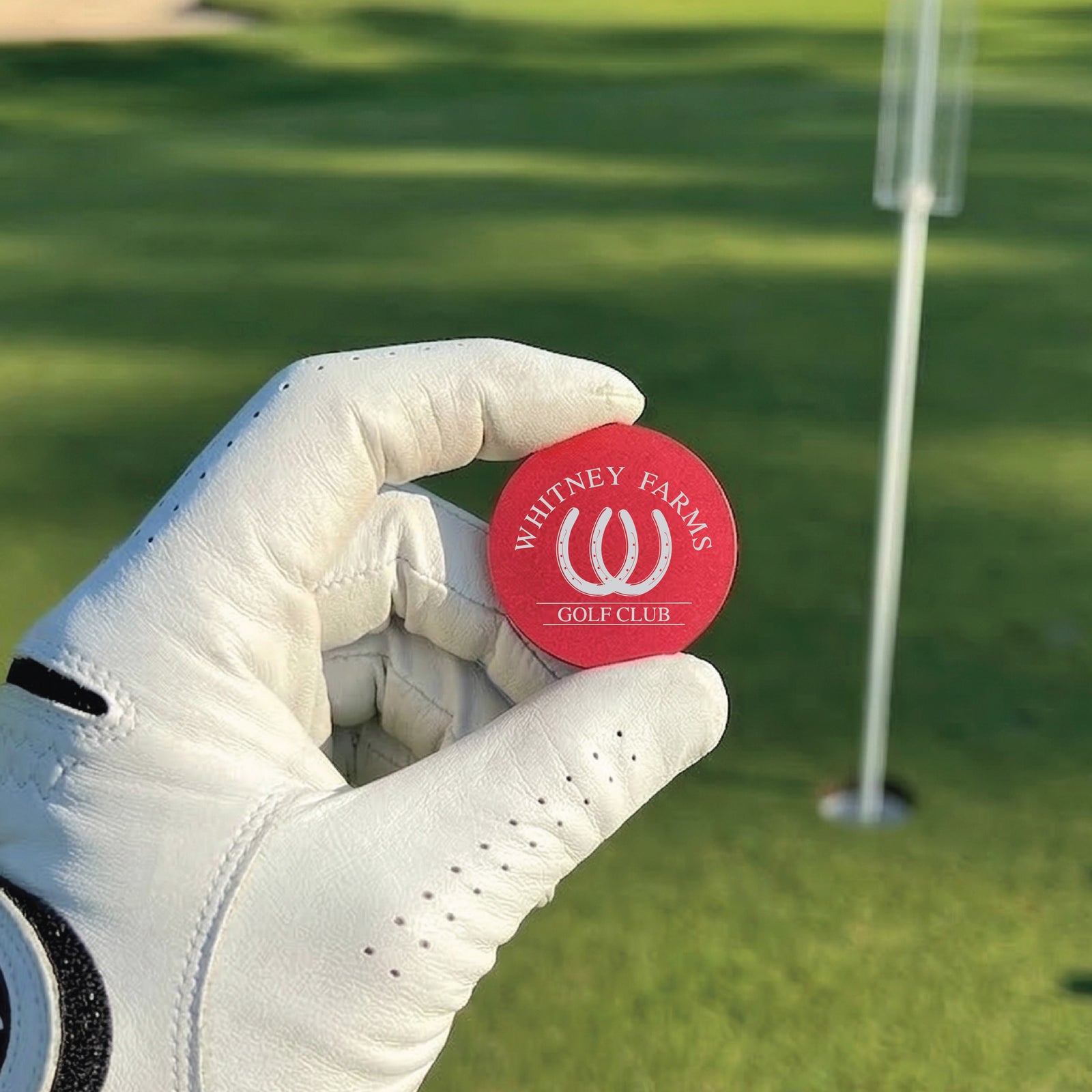 Red ball marker held in gloved hand, engraved with sample "Whitney Farms Golf Club" logo and text, golf course green and flagstick blurred in background