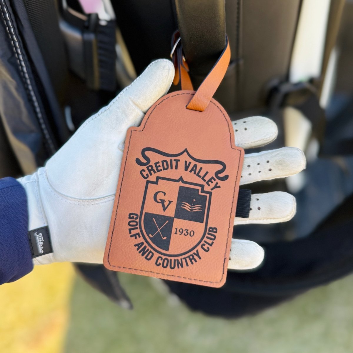 Tan vegan leather bag tag with engraved "Credit Valley Golf and Country Club" logo and "1930" text, displayed in golfer's hand wearing white glove, with three tees inserted in slots