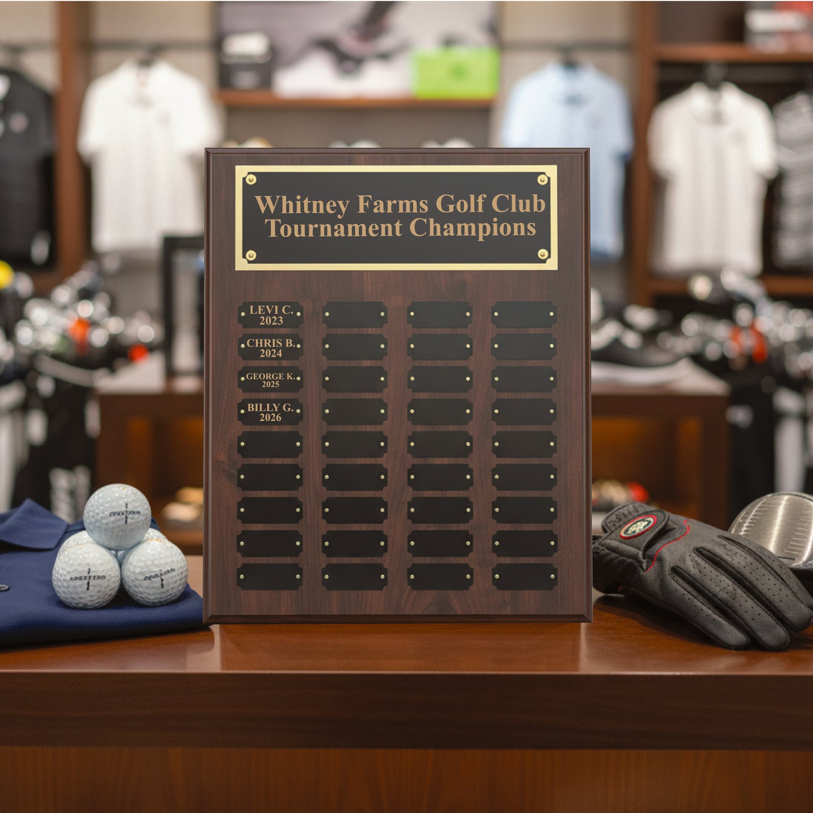 Dark wood perpetual plaque with engraved brass header reading "Whitney Farms Golf Club Tournament Champions" and sample engraved names including "LEVI C. 2023," "CHRIS B. 2024," "GEORGE K. 2025," and "BILLY G. 2026" with multiple blank nameplates for future winners.