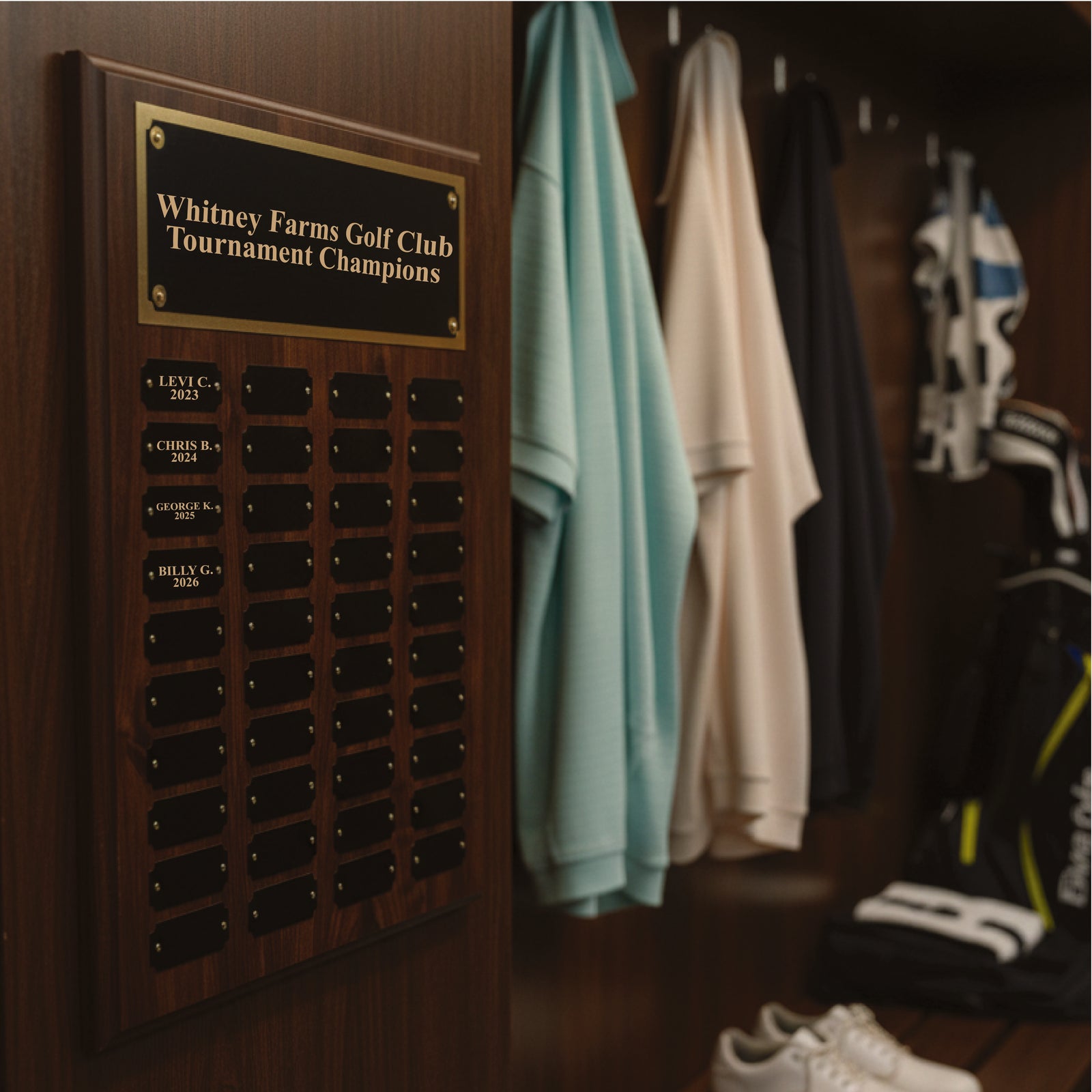 Wood plaque with engraved header reading "Whitney Farms Golf Club Tournament Champions" displaying multiple rows of black name plates with sample champion names and years from 2023 to 2026, mounted on clubhouse wall near hanging golf towels.