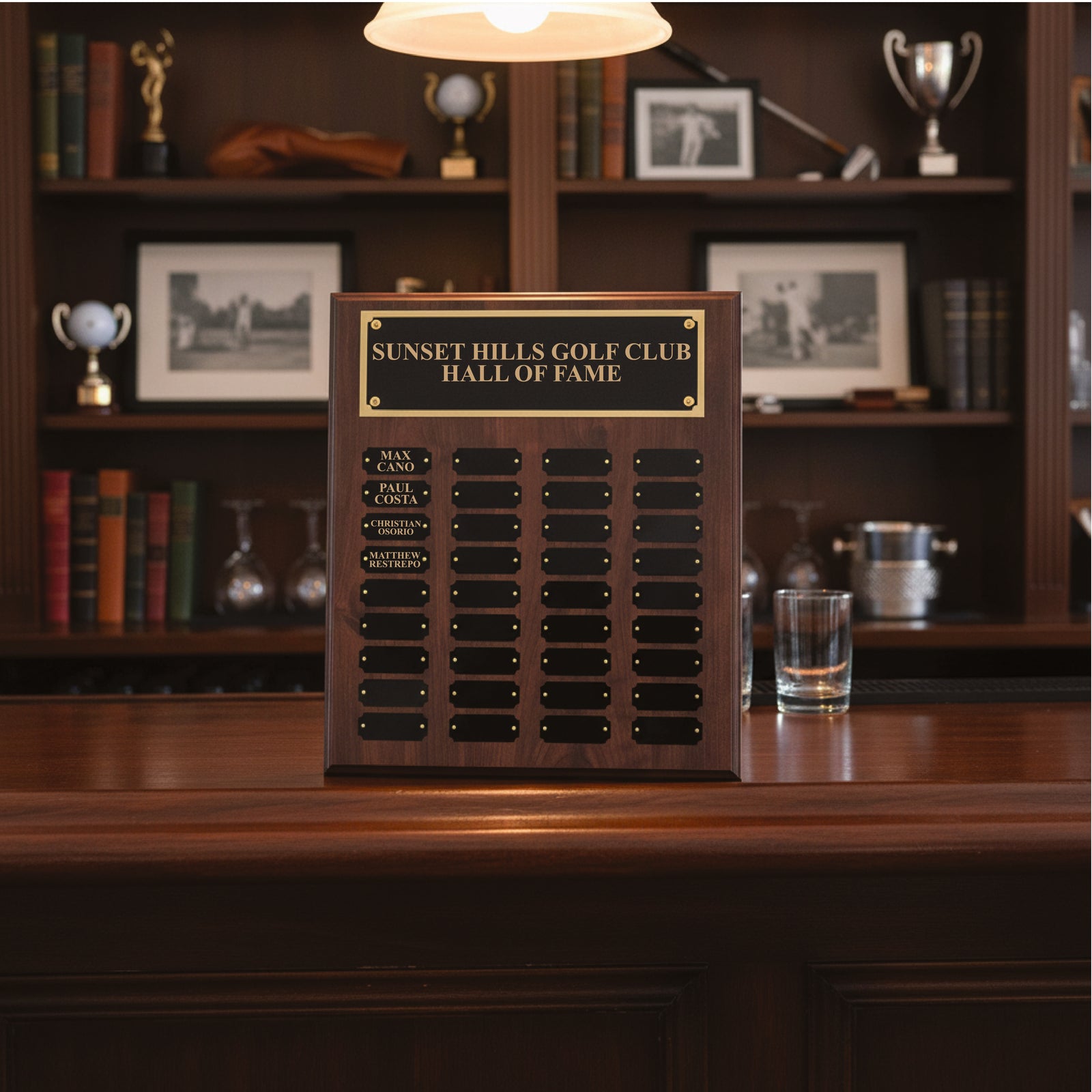 Dark wood perpetual plaque with gold-engraved header reading "Sunset Hills Golf Club Hall of Fame" and multiple rows of black nameplates mounted on wooden base, displayed on desk in office setting.