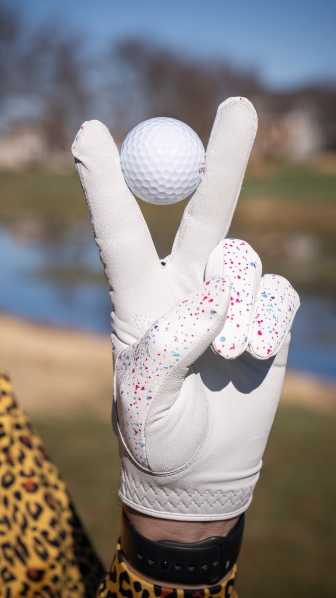 White Cabretta leather golf glove with multicolored paint splatter design, displayed on hand holding white golf ball on course.