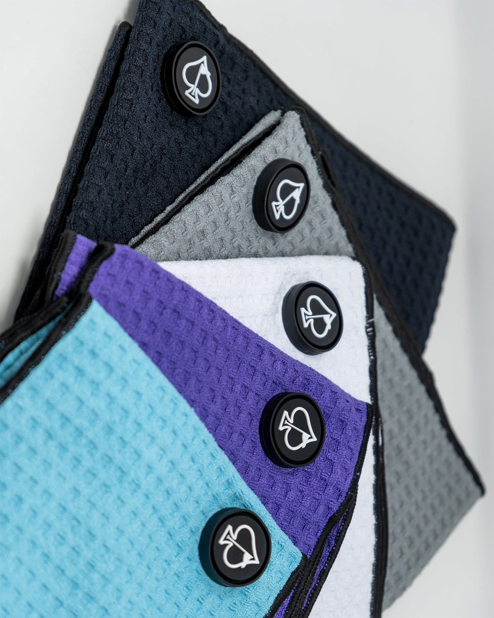 Stack of five golf towels in navy, gray, white, purple, and sky blue, each with a black circular magnetic clip featuring a white spade logo