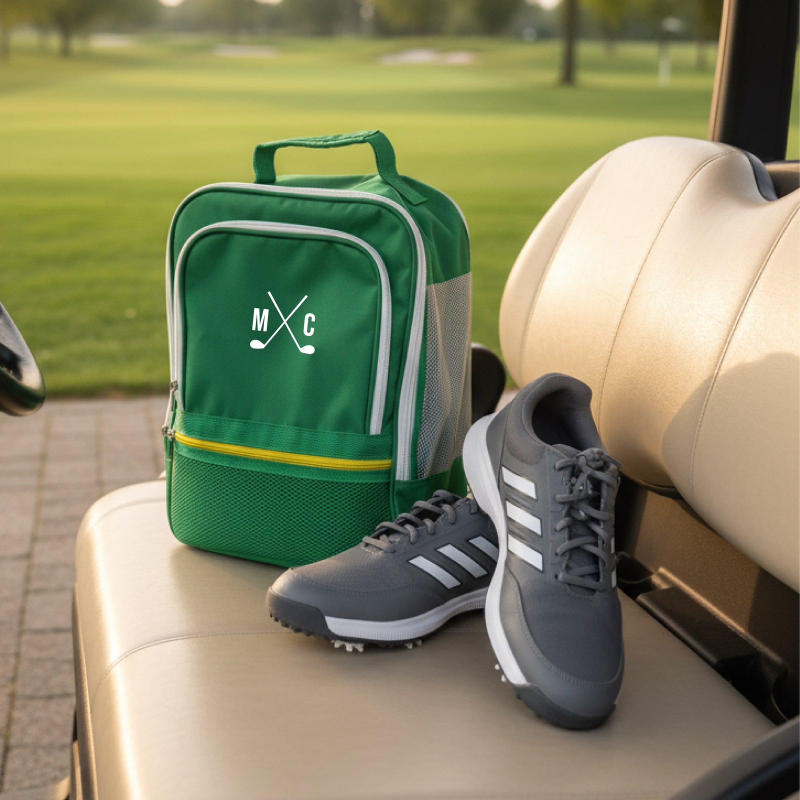 Green golf shoe bag with embroidered initials "MC" and crossed clubs design, displayed on golf cart seat next to black and white golf shoes