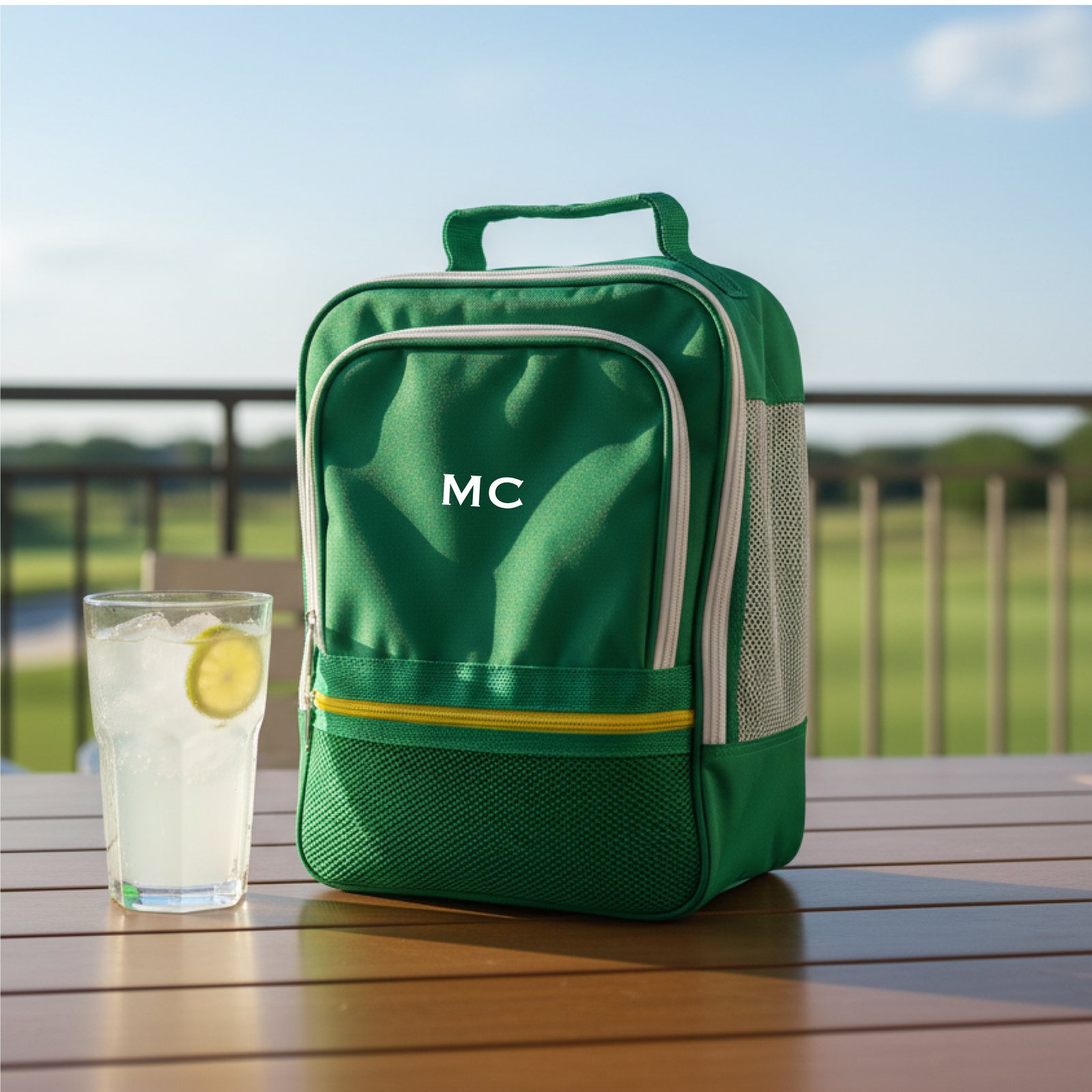 Green golf shoe bag with sample initials "MC" embroidered on front, displayed on outdoor wooden table next to glass of lemon water with golf course in background