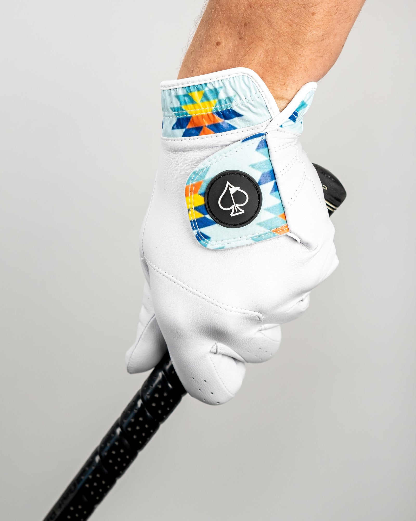 White golf glove with Southwest geometric pattern on cuff, featuring magnetic ball marker with spade symbol, worn on hand holding black golf club grip.