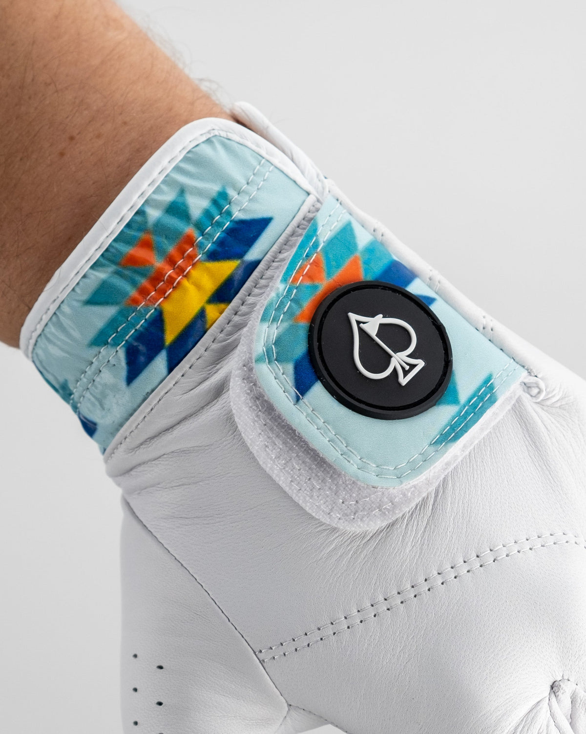 White leather golf glove with turquoise cuff featuring geometric diamond pattern in orange, blue, and yellow, black ball marker with spade icon visible on wrist.