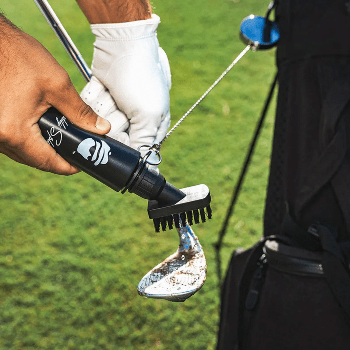 Golfer in white glove holding black golf club brush with retractor clip, cleaning club head on green course with dirt visible on brush bristles