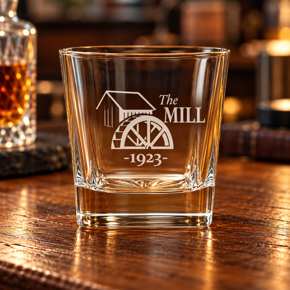 Clear rocks glass etched with "The MILL" logo featuring a watermill graphic and "-1023-" below, filled with amber whiskey, on wooden table with blurred bar background.