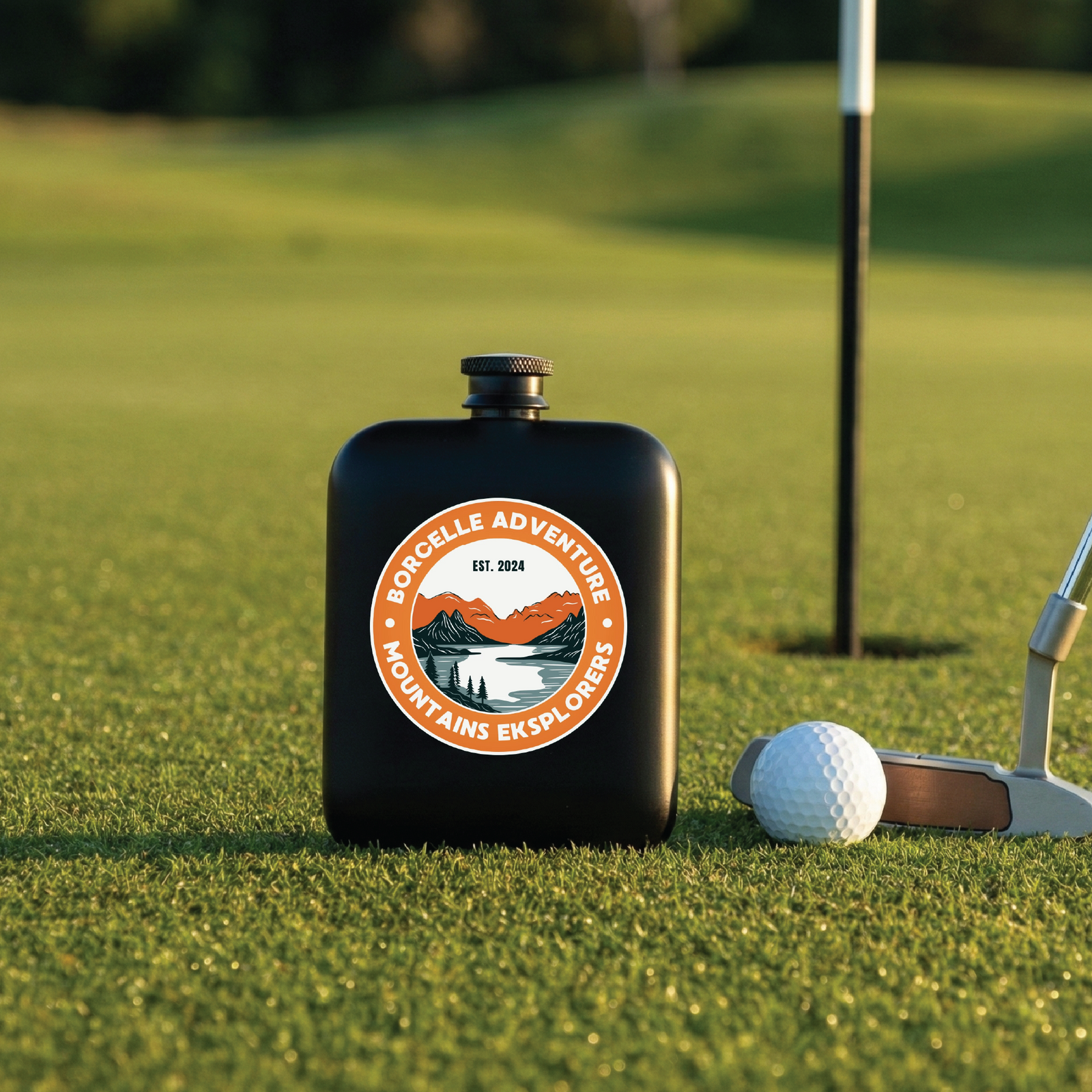 Black stainless steel flask with orange and white circular logo reading "Borcelle Adventure Mountains Eksploreers Est. 2024" displayed on putting green next to golf ball and putter.