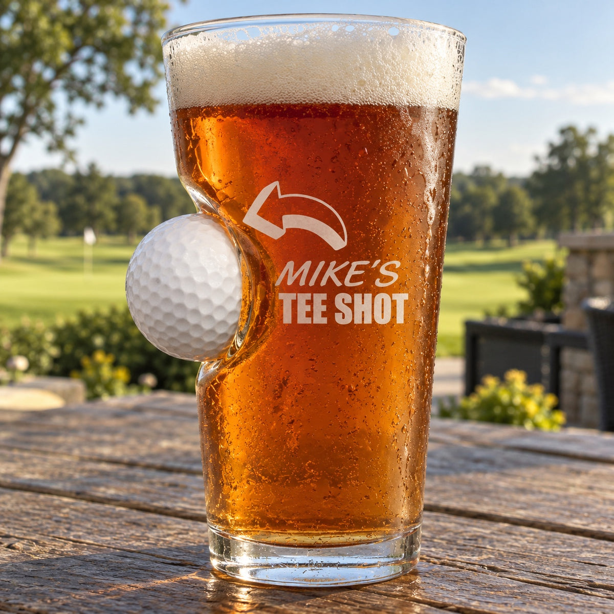 Golf pint glass full of amber ale with a real golf ball set into the side, printed with an arrow and MIKE'S TEE SHOT, on a clubhouse patio