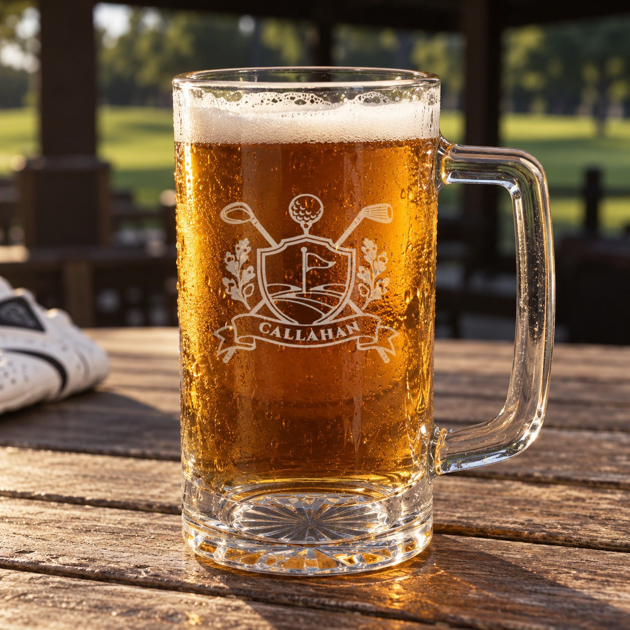 Golf crest beer mug engraved CALLAHAN, filled with amber ale on a clubhouse patio table with the golf course behind it