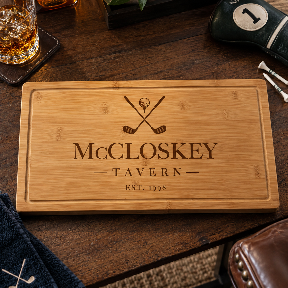 Bamboo cutting board engraved "McCLOSKEY TAVERN" with crossed golf clubs design and "EST 1998" at the bottom, displayed on a dark wood table with golf accessories nearby