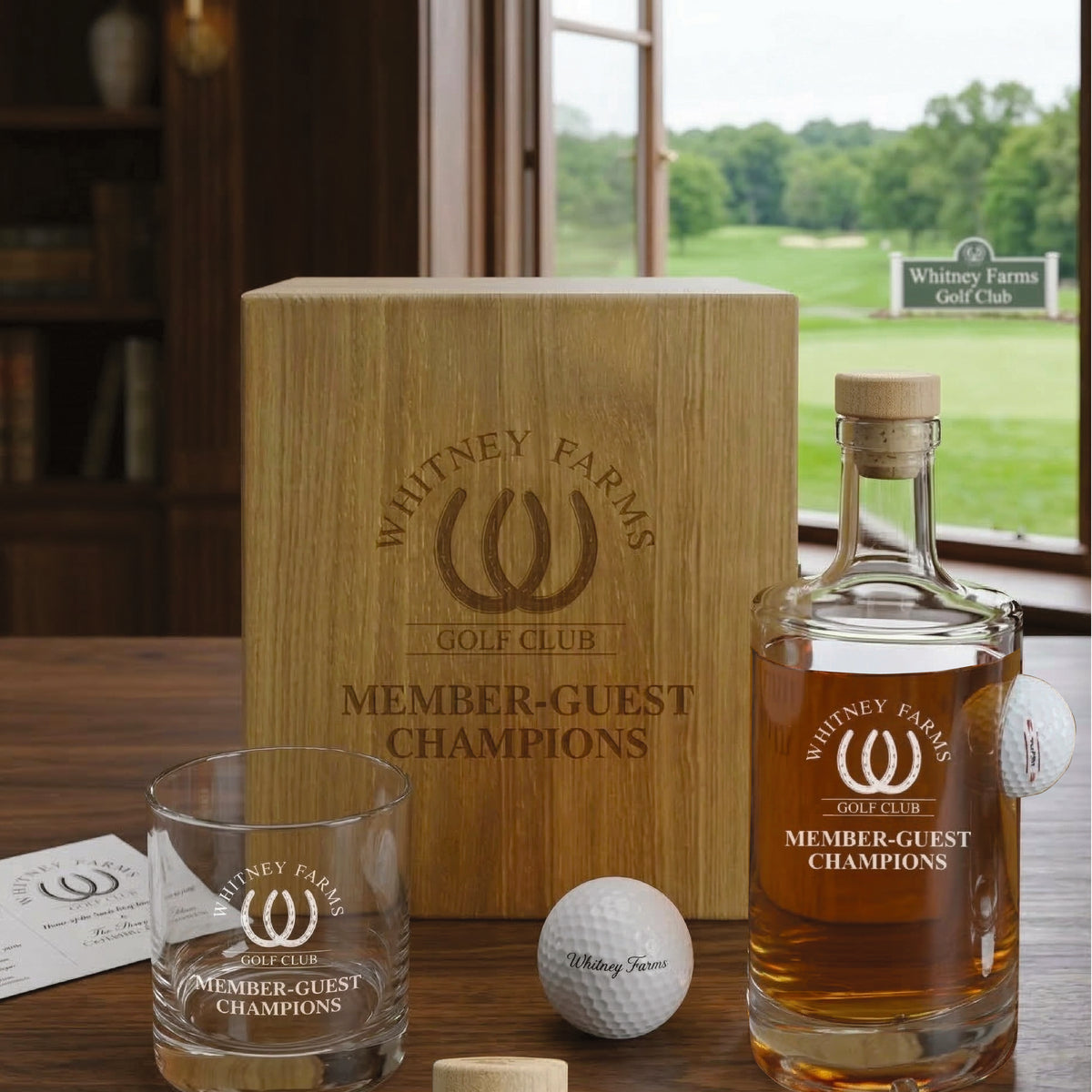 Wooden box and decanter set engraved with Whitney Farms Golf Club logo and "Member-Guest Champions," displayed with matching whiskey glass and golf ball on wooden table by window overlooking golf course