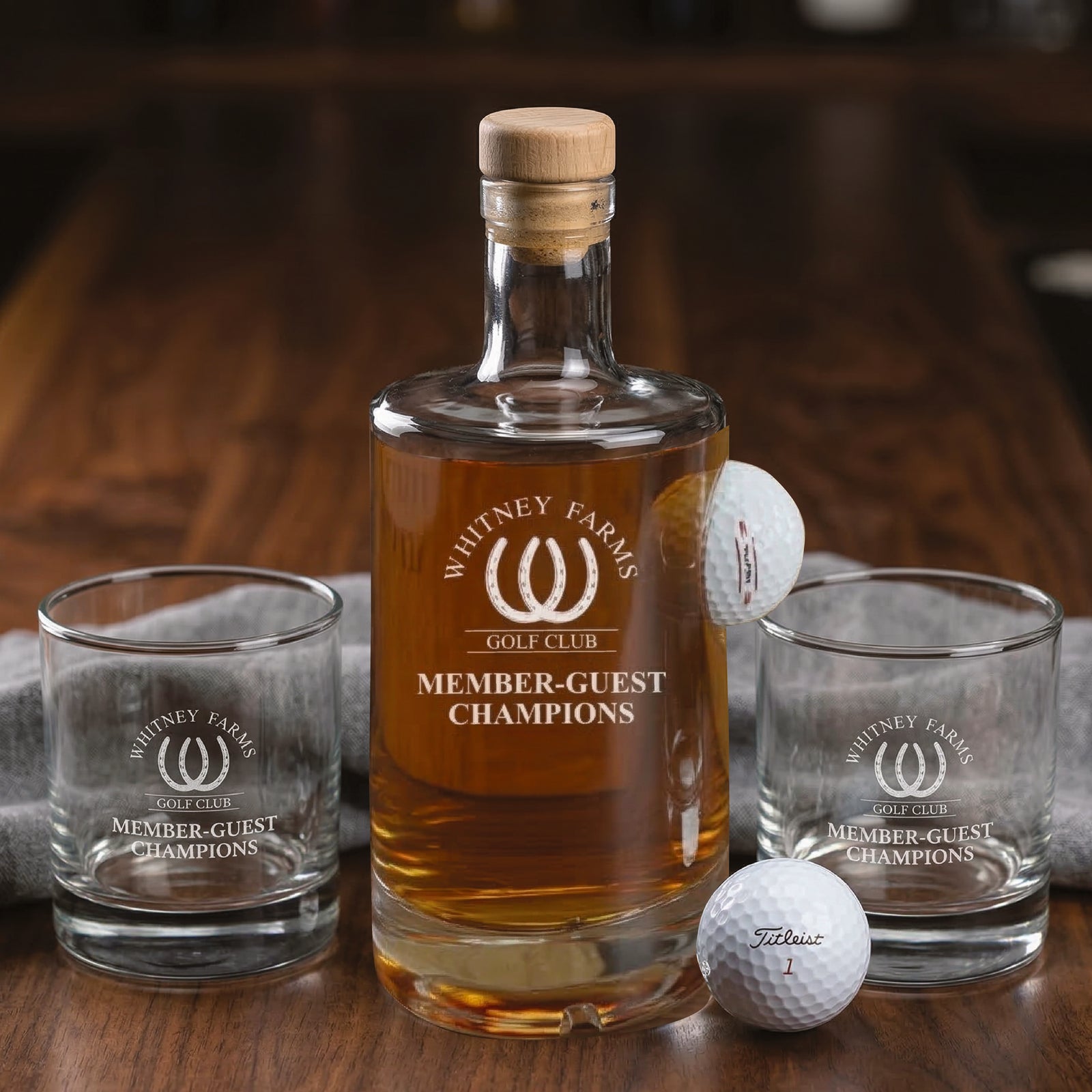 Golf-themed decanter set with wooden stopper filled with amber spirits, two matching rocks glasses, and two white golf balls, all engraved with "Whitney Farms Golf Club Member-Guest Champions" logo
