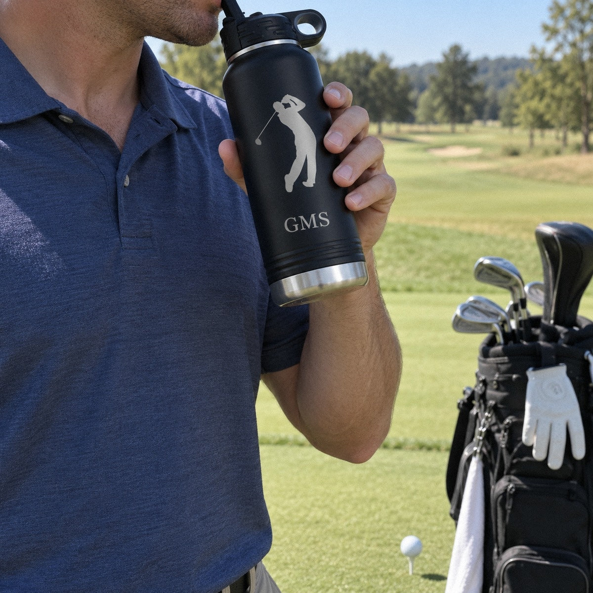 A golfer drinks from his personalized water bottle on the tee box beside his golf bag, the bottle engraved with a golfer and GMS
