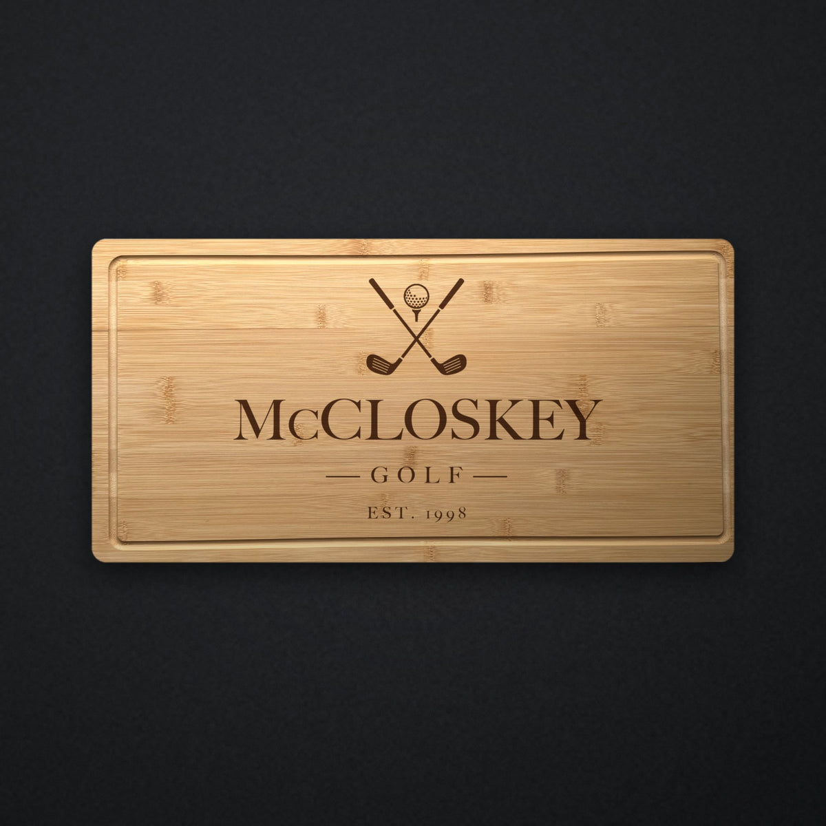 Personalized golf cutting board in solid bamboo on dark slate, engraved with crossed golf clubs and a ball above the family name and the year.
