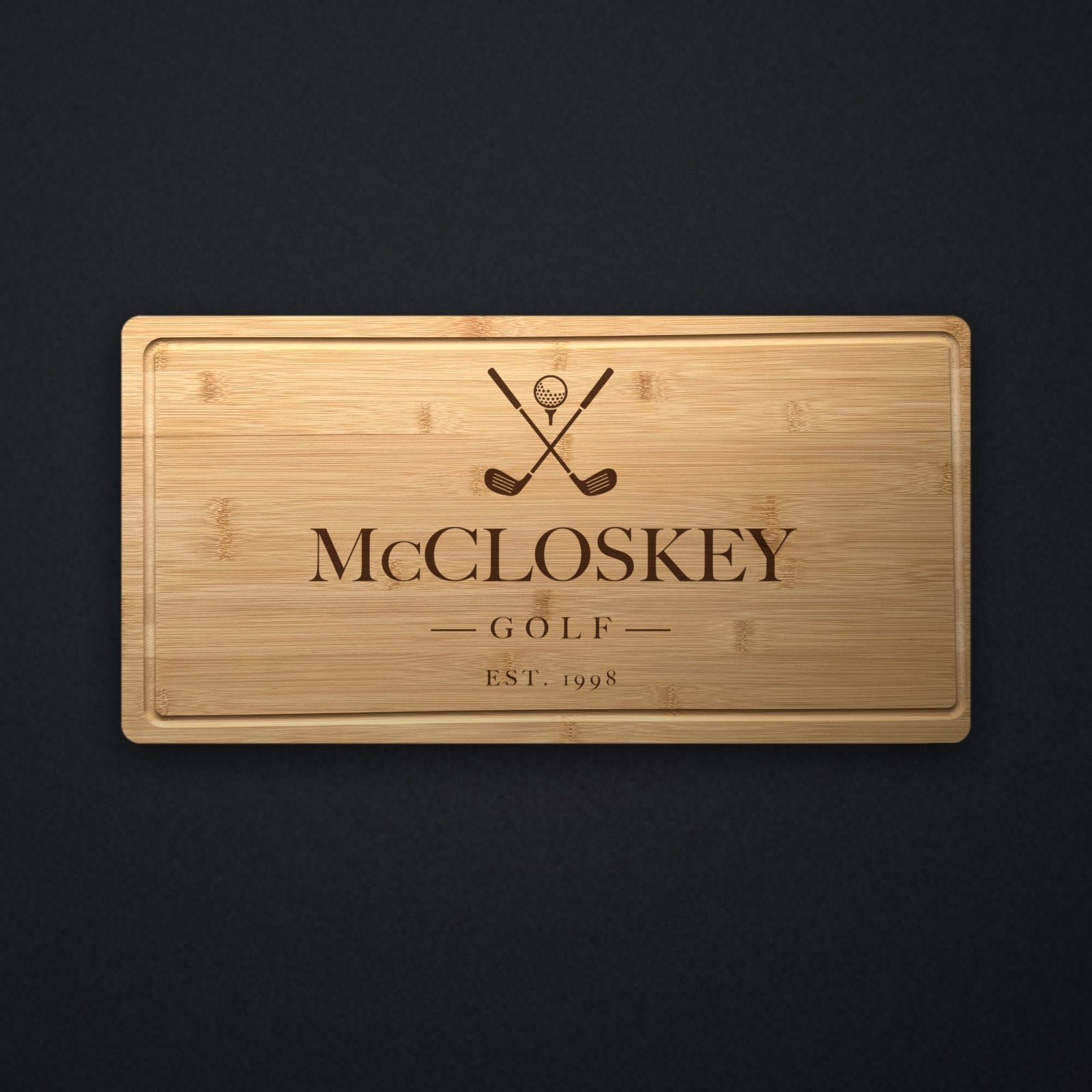 Personalized golf cutting board in solid bamboo on dark slate, engraved with crossed golf clubs and a ball above the family name and the year.