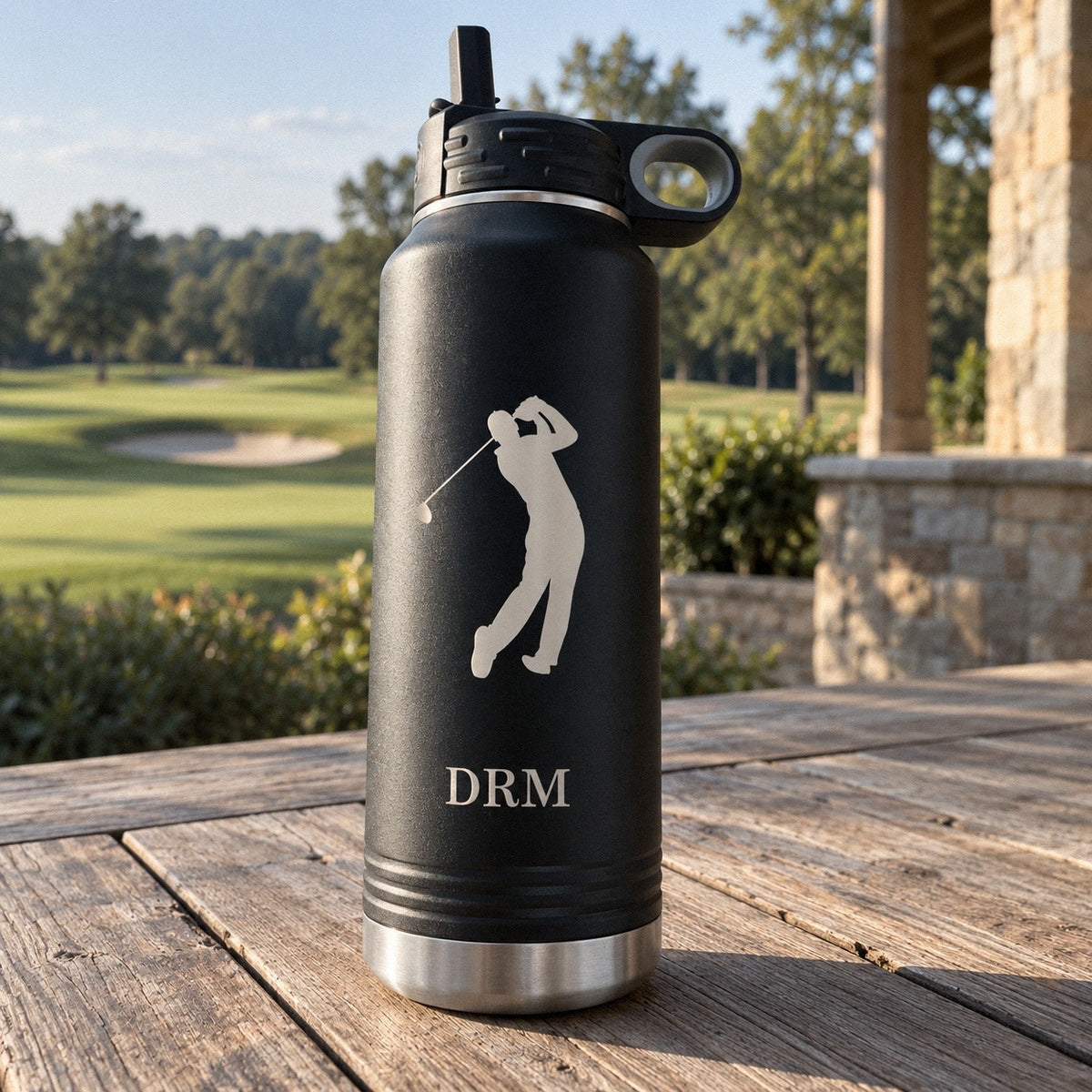 Personalized golf water bottle in matte black on a clubhouse patio table, engraved with a golfer silhouette and the initials DRM