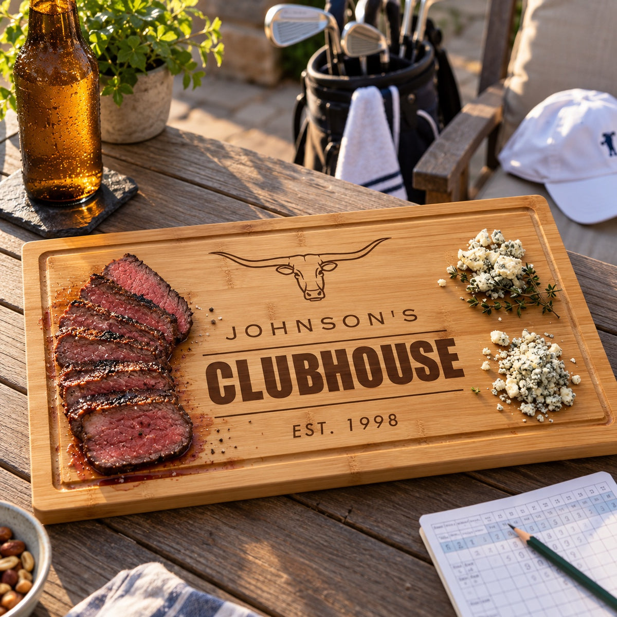 Personalized steakhouse cutting board engraved Johnson's Clubhouse Est. 1998 with sliced steak on a patio table beside a golf bag, a gift for a golfer who loves steak