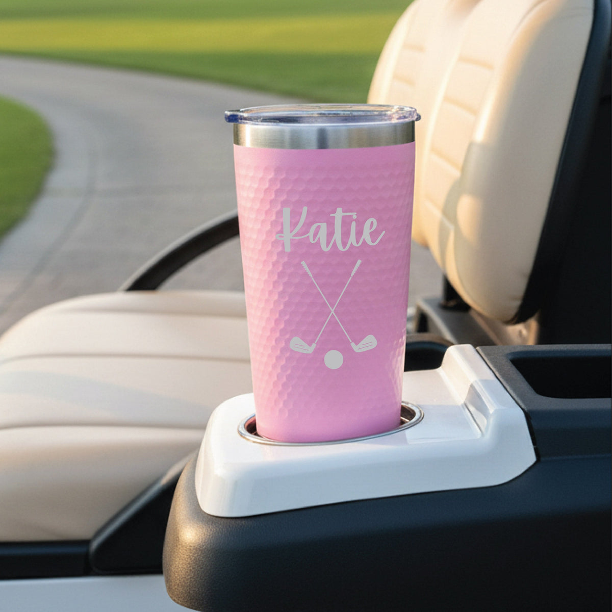 Pink insulated tumbler with golf ball texture, engraved with sample name "Katie" and crossed golf clubs design, sitting in car cup holder with golf course visible in background