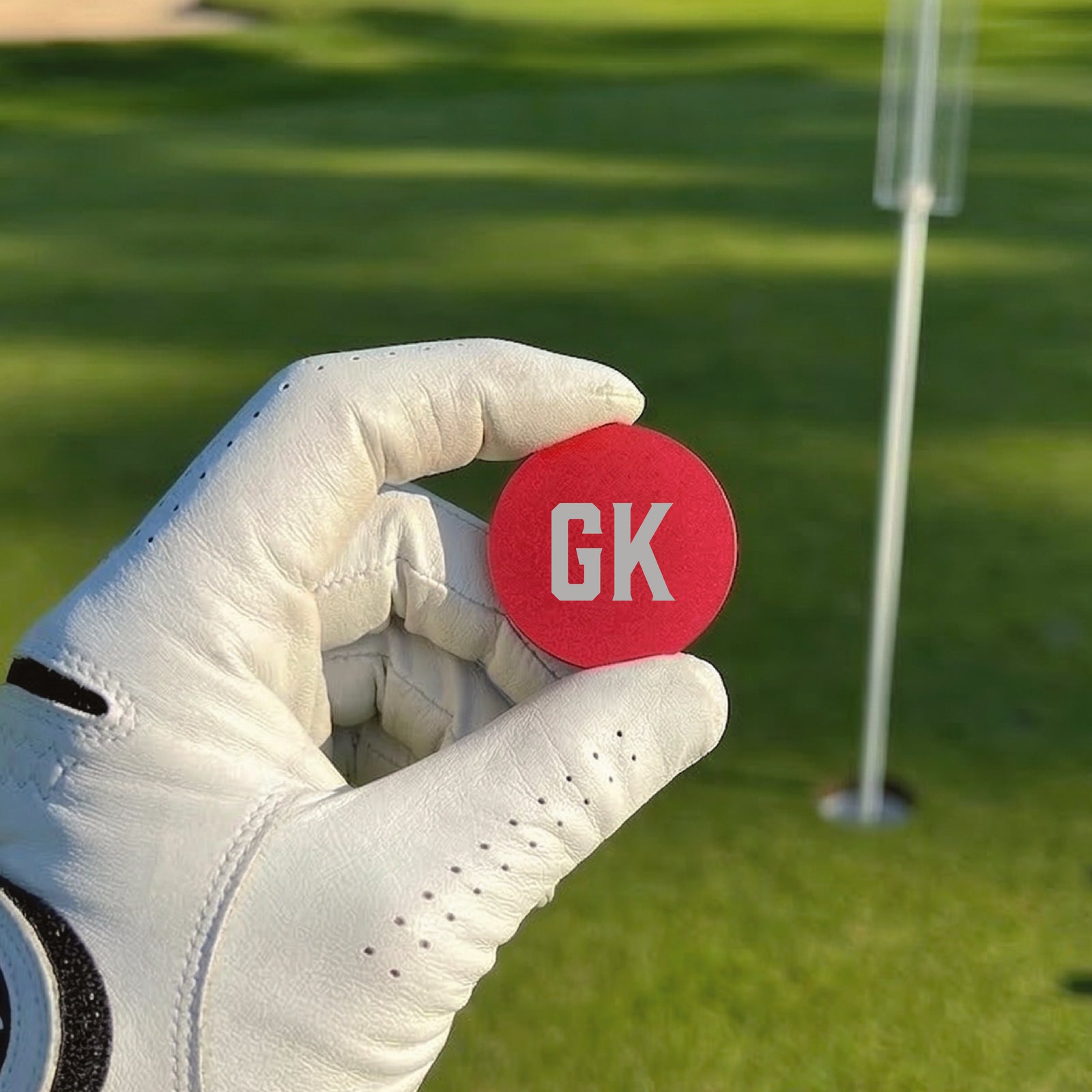 Red ball marker with sample initials "GK" in white, held in a gloved hand above a golf green with flagstick visible in background