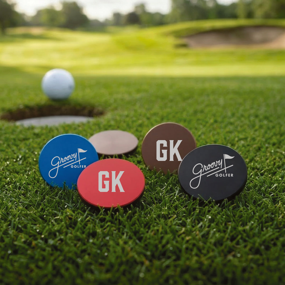 Four personalized ball markers in blue, red, brown, and black displayed on golf course grass with a golf ball and hole in background