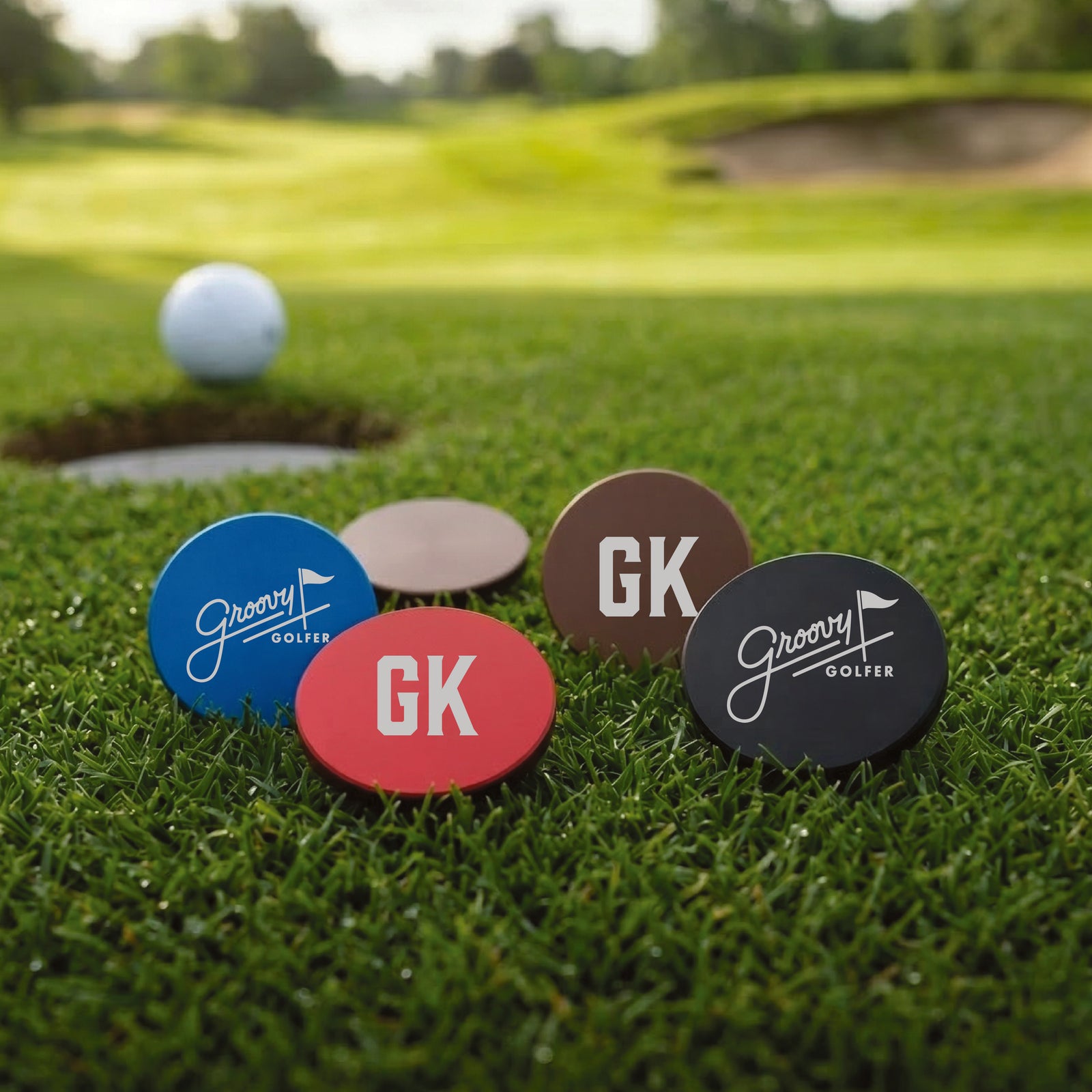 Four personalized ball markers in blue, red, brown, and black displayed on golf course grass with a golf ball and hole in background