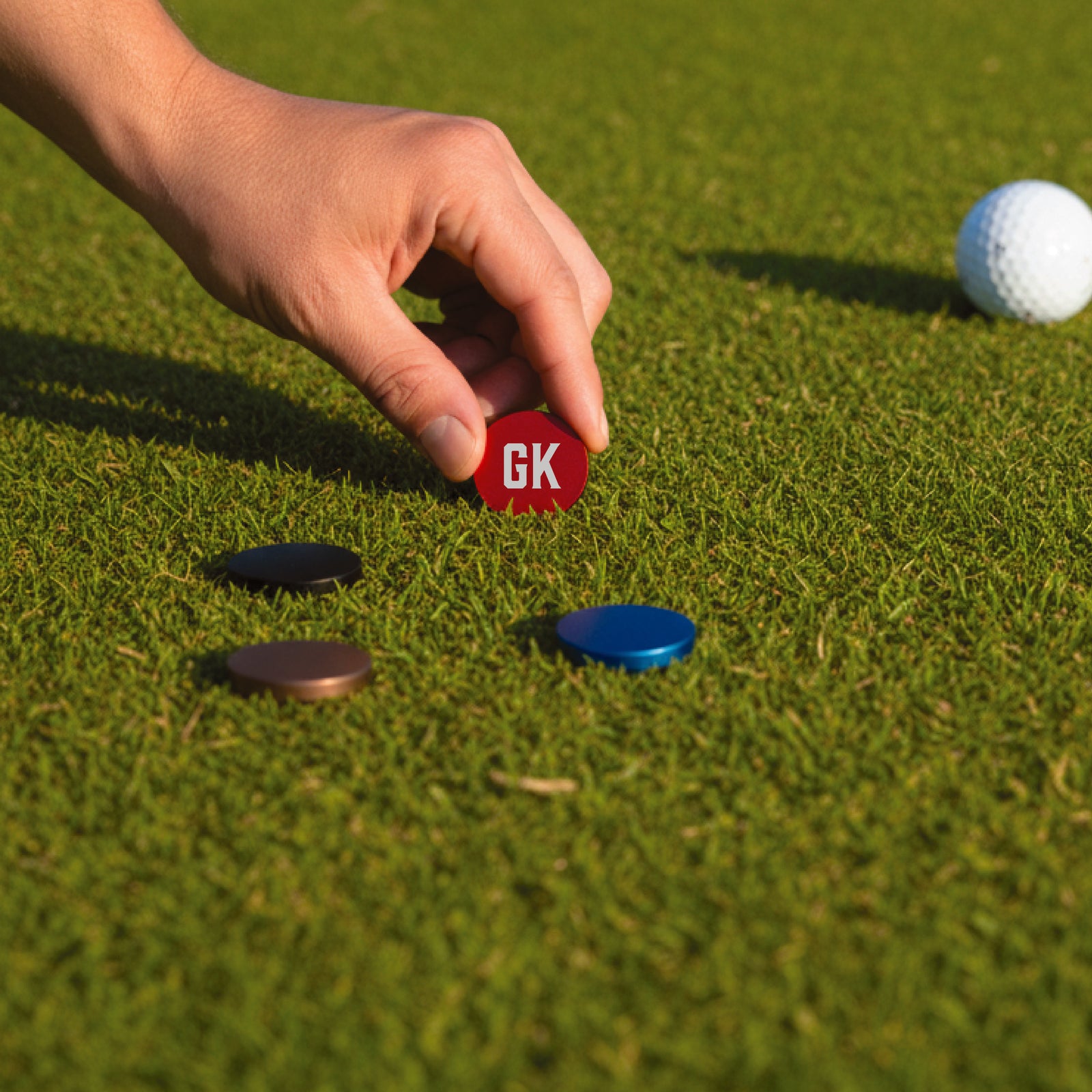 Hand placing a red ball marker engraved with sample initials "GK" on putting green near golf ball and other colored markers