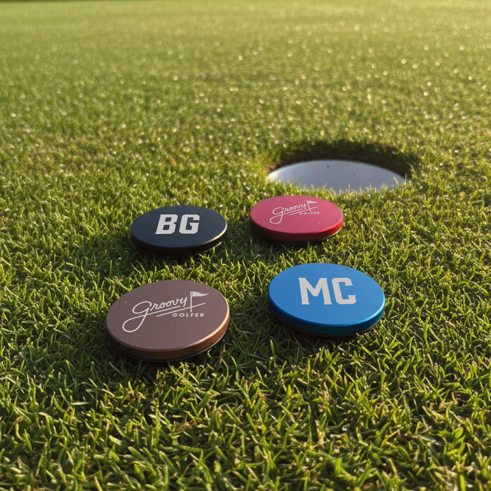 Four oval ball markers on golf green with sample personalization: black marker engraved "BG", brown marker with Groovy Golfer logo, pink marker engraved "BG", and blue marker engraved "MG