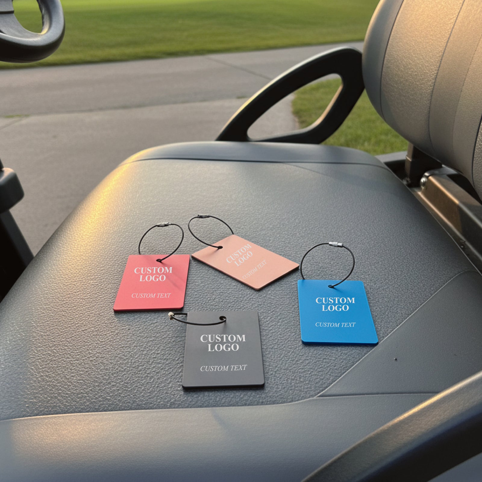 Four aluminum golf bag tags in red, peach, gray, and blue displayed on a car dashboard, each showing sample "Custom Logo" and "Custom Text" engraving with steel cable loops