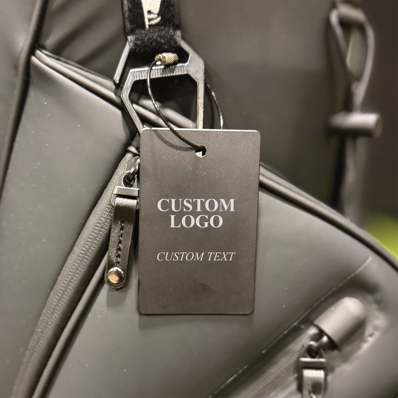 Black aluminum golf bag tag attached to gray golf bag with steel cable loop, displaying sample custom logo and text areas for personalization