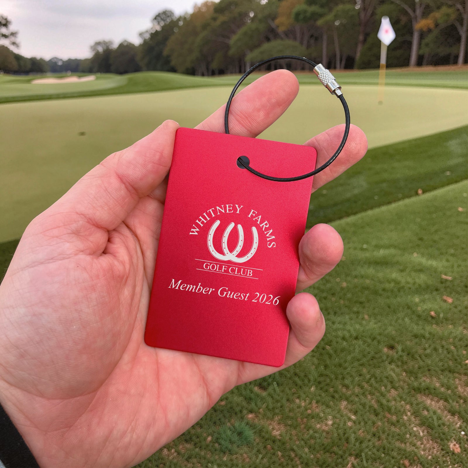 Red aluminum bag tag held in hand on golf course, engraved with sample text "Whitney Farms Golf Club" and "Member Guest 2026" with black steel cable loop
