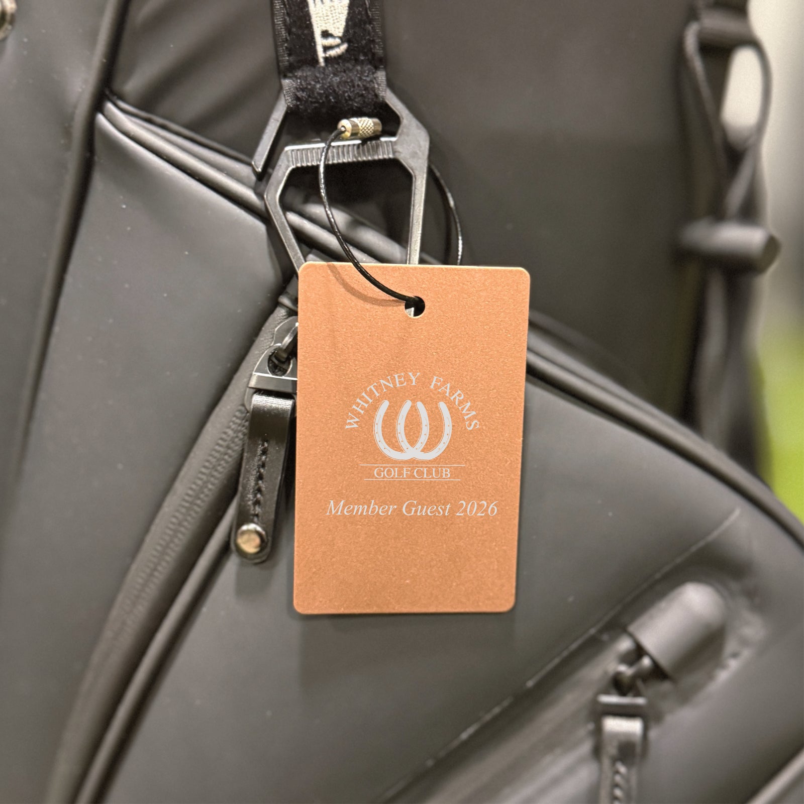 Peach-colored aluminum bag tag engraved with sample golf club logo and "Member Guest 2026" text, attached to black golf bag with steel cable loop