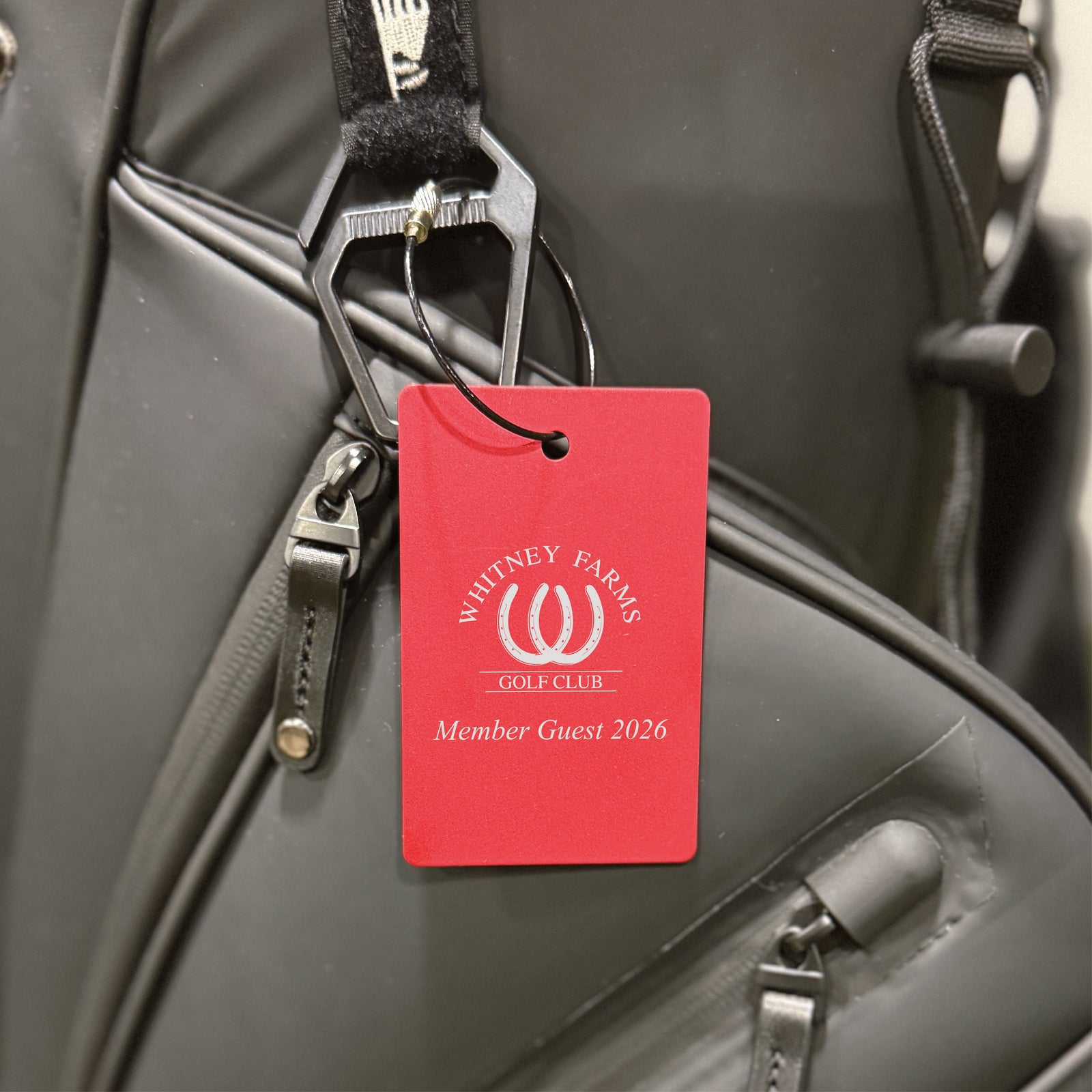 Red aluminum golf bag tag engraved with "Whitney Farms Golf Club" logo and "Member Guest 2026" text, attached to black golf bag with steel cable loop