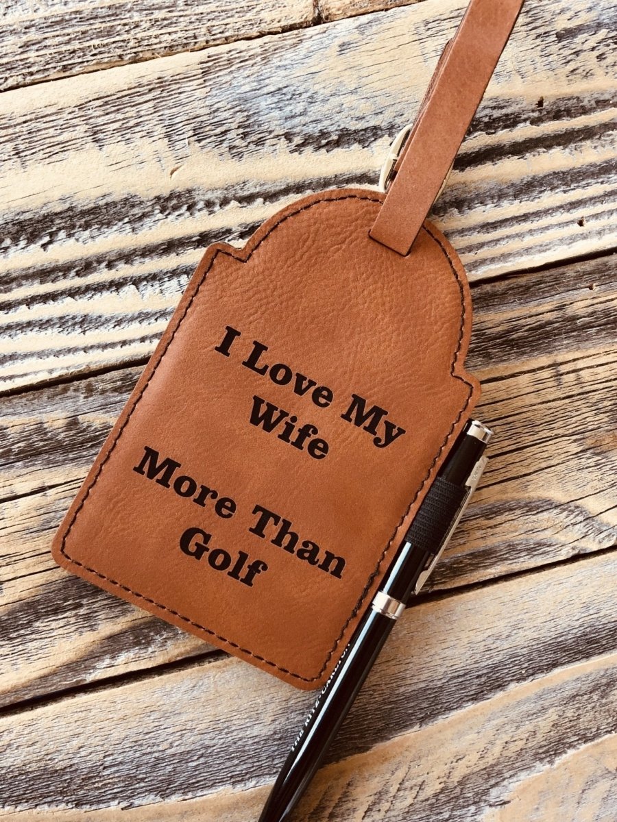 Tan leather golf bag tag with stitched edges, engraved with sample text "I love My Wife More Than Golf" and attached to golf club handle with leather strap