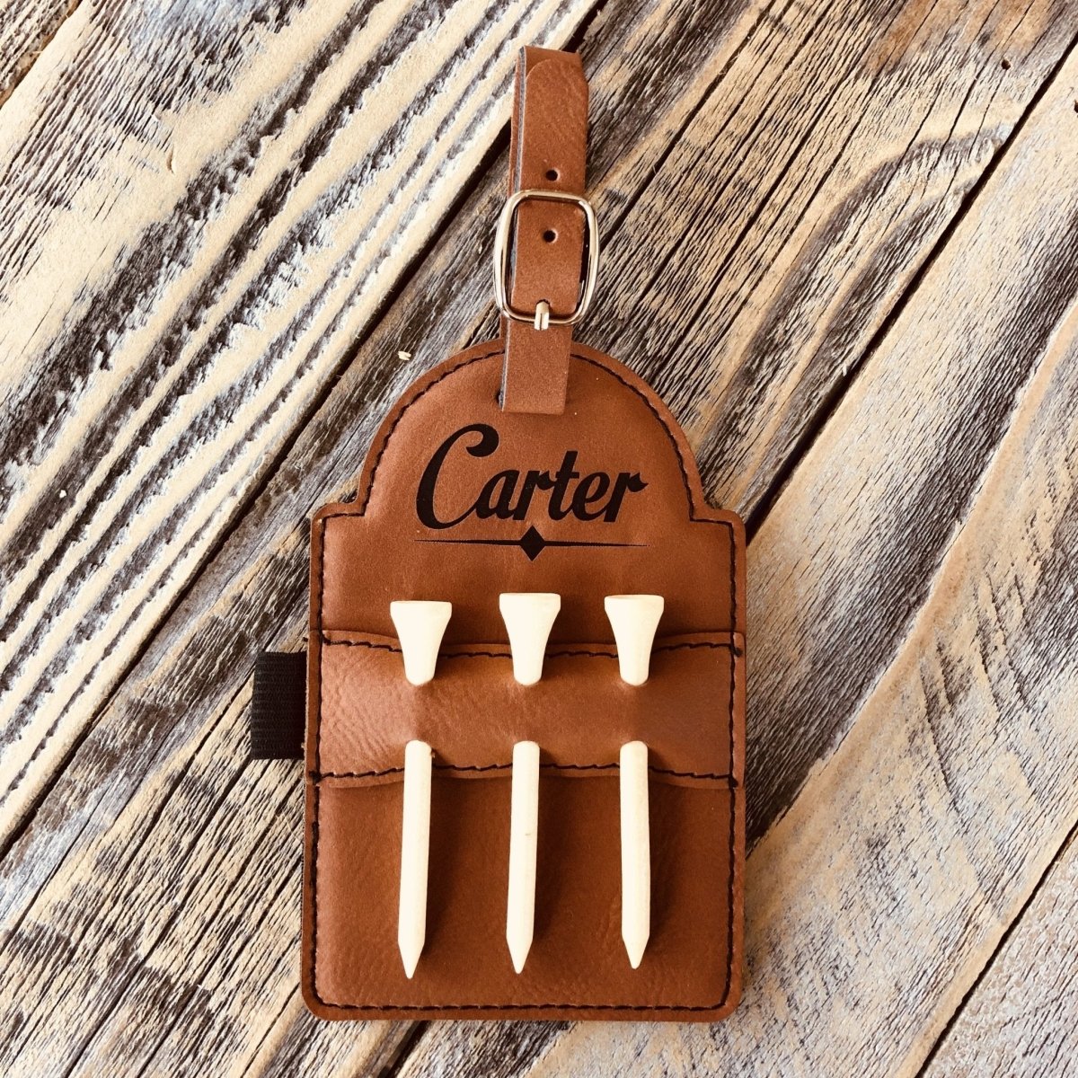 Tan leather golf bag tag with sample name "Carter" engraved at top, holding six white wooden golf tees, attached with brown leather strap and metal buckle